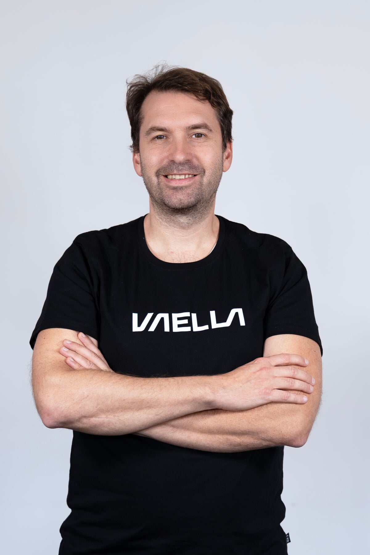 A man smiles confidently in a black Vella-branded t-shirt.