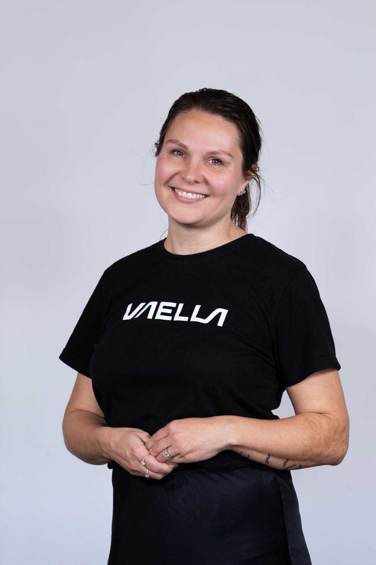 A woman smiles, wearing a black VAELLA t-shirt with visible rings on her fingers.